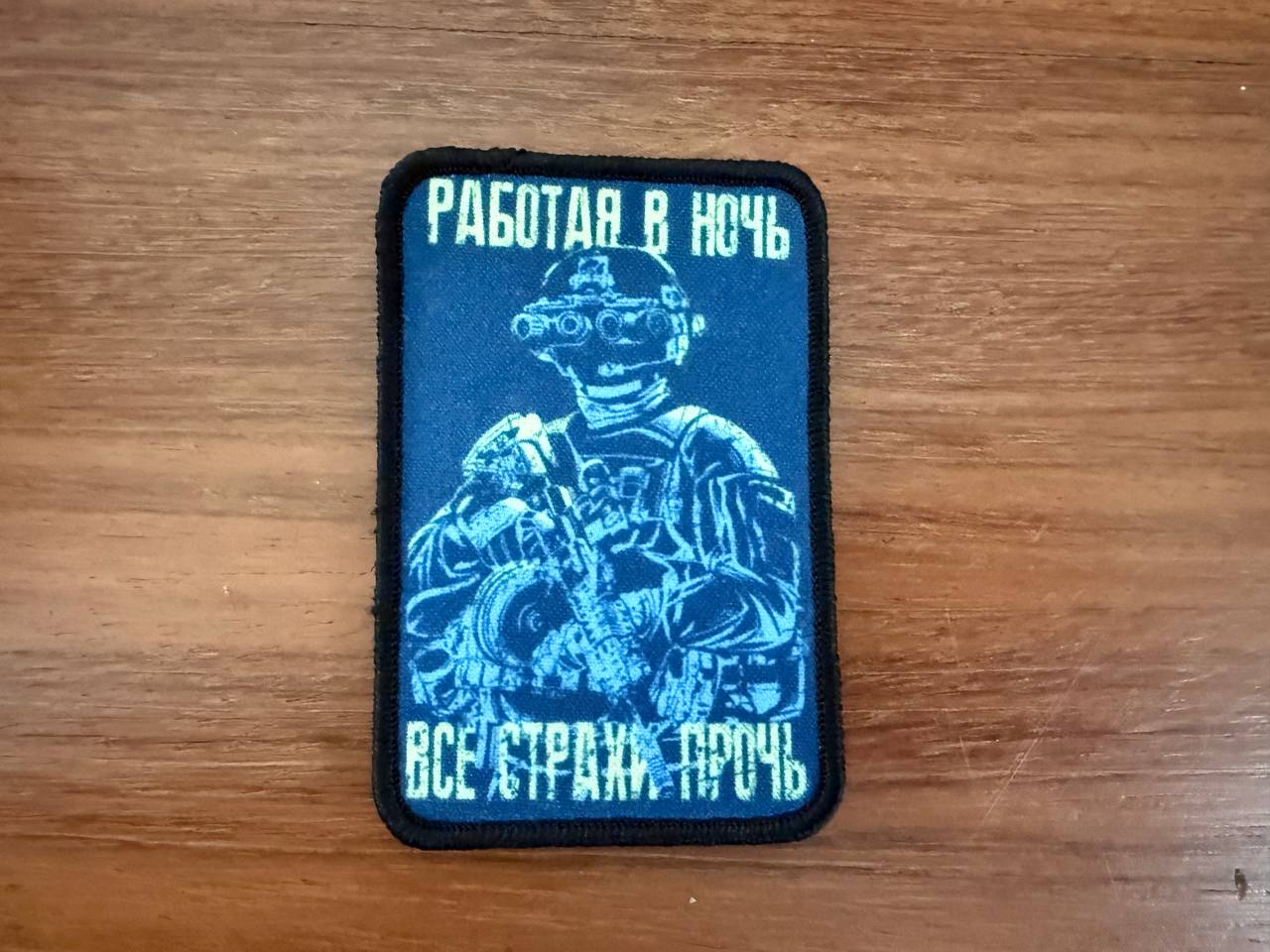 Blue patch with a person wearing night-vision glasses  and text on a wooden surface