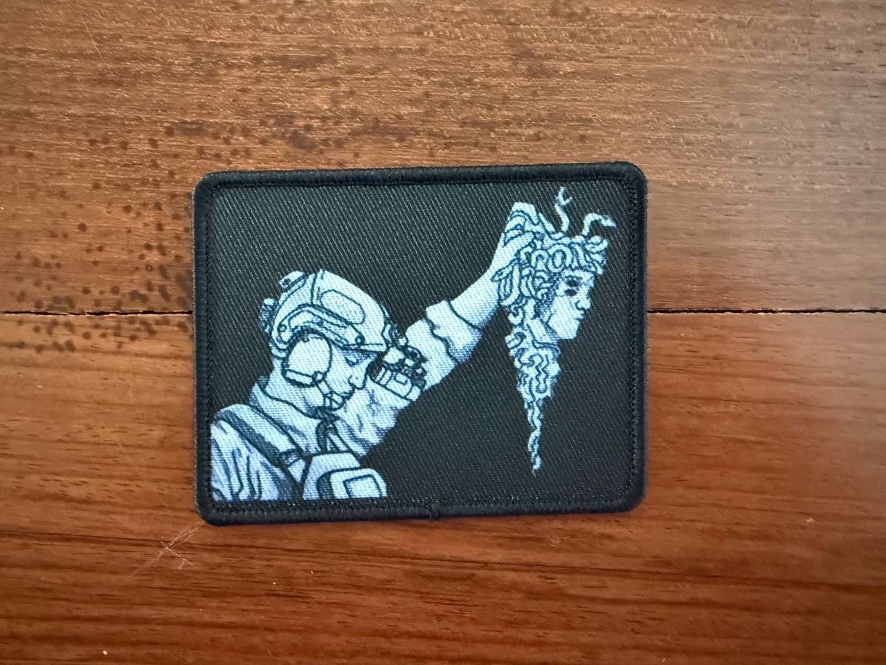 Patch with a design of a person in a helmet holding a decapitated Medusa's head with snakes