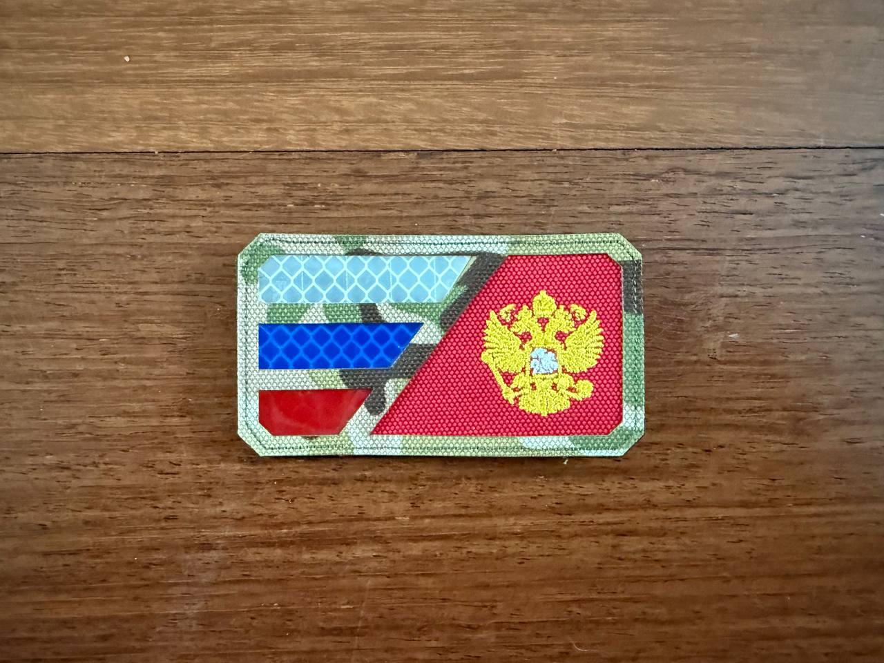The Russian Tricolor Flag and Emblem Design Patch - Military Badge by Tanki Store features two blue stripes, a red stripe, and a gold double-headed eagle on red. Ideal for buying patches in bulk. Patch shown on a wooden surface.