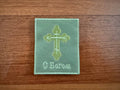 A green rectangular fabric patch from Tanki Store’s set, featuring an embroidered gold Orthodox cross and Cyrillic “С Богом” (May God be with you), from the Russian Orthodox Cross Patch May God be with you badge. Placed on wood.