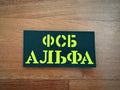 A green and yellow Russian FSB Alpha Unit Patch (15x7.5cm) from Tanki Store lies on a wooden surface—just one of many military badges available in large quantity at the best price.
