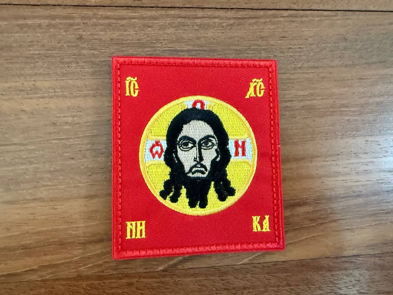 Embroidered patch with a central figure on a red background with symbols around, on a wooden surface.