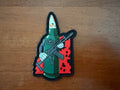 Patch of a green character holding a rifle on a wooden surface