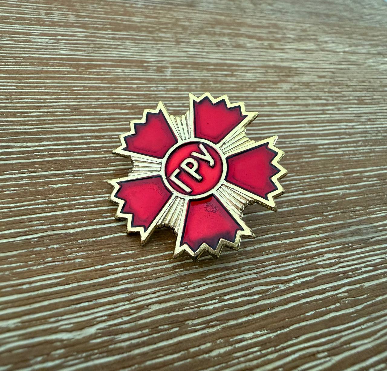 Russian GRU Service Pin featuring a red and gold star-shaped design with Cyrillic letters, displayed on a textured wooden surface, vintage Russian surplus style.