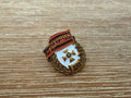 Russian Donetsk People's Republic (DNR) Pin featuring a laurel wreath, a central gold cross emblem, and a red and black banner with Cyrillic text on a wooden surface.