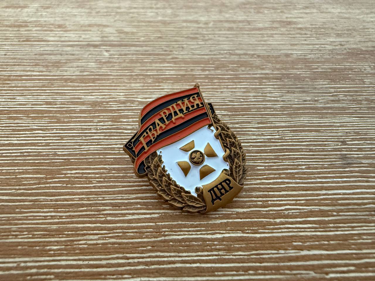 Russian Donetsk People's Republic (DNR) Pin featuring a laurel wreath, a central gold cross emblem, and a red and black banner with Cyrillic text on a wooden surface.