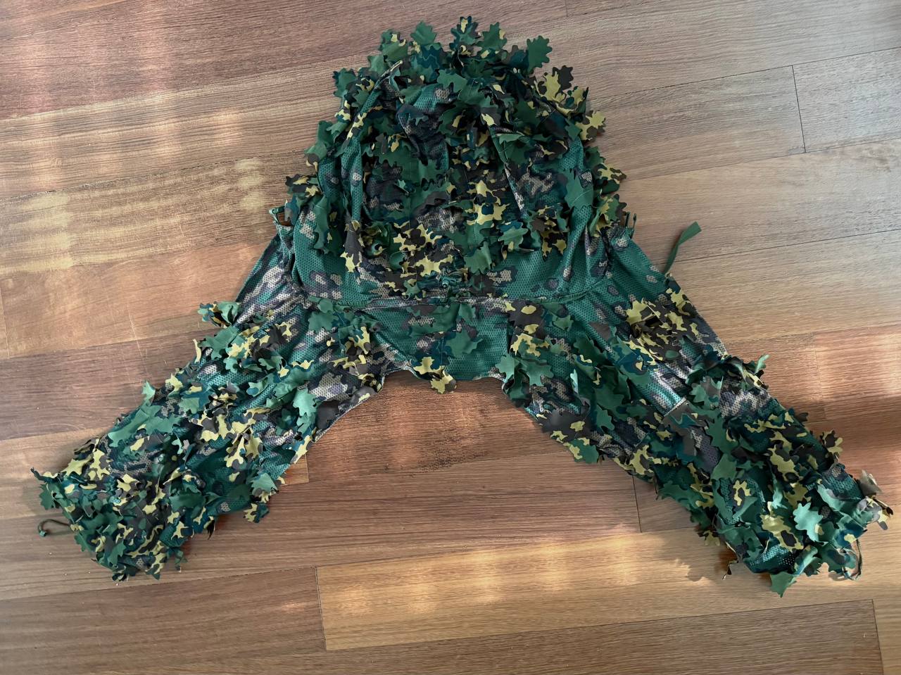 Russian SS-Leto Leaf Cape / Viper Hood laid flat on a wooden floor, featuring green and brown camouflage fabric with leaf-like attachments for effective concealment.