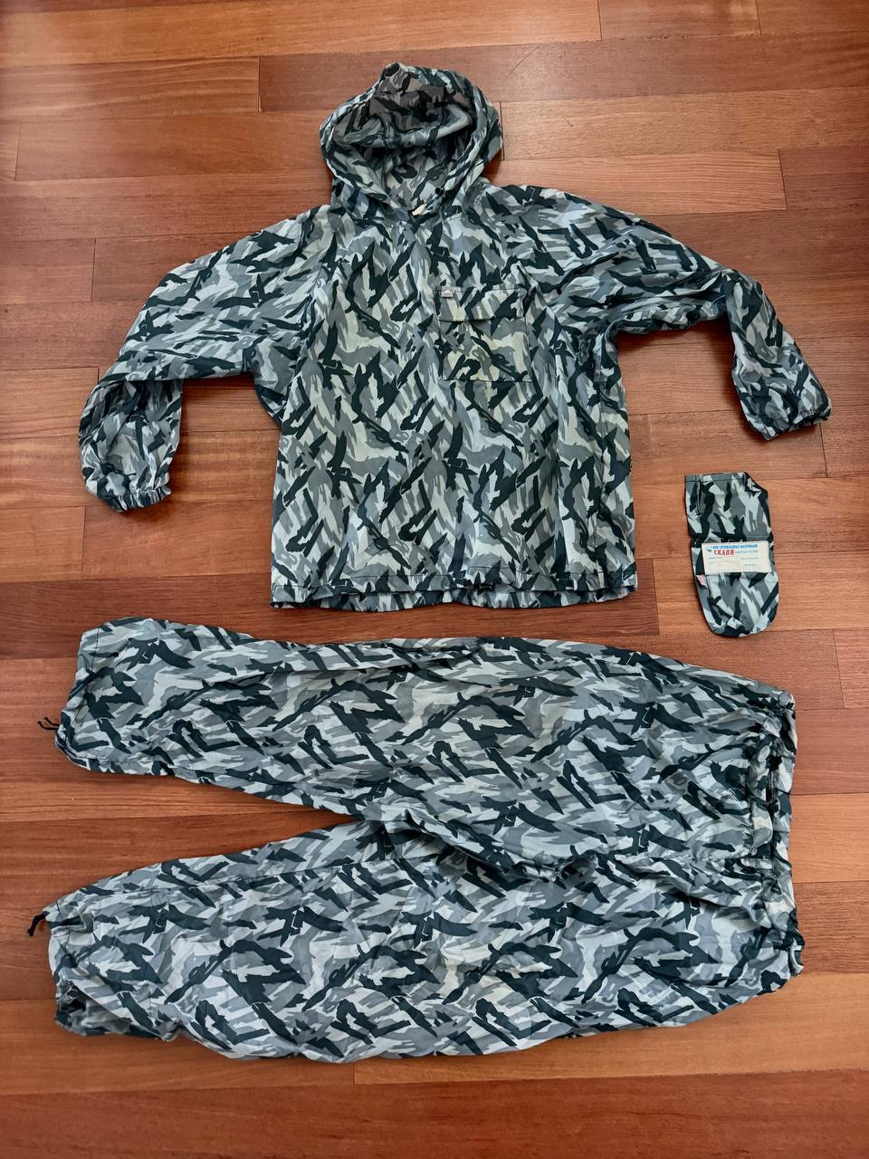 Russian NPO-SM "Sklon" Water-resistant/Windproof Suit – Melted Snow Color laid flat on a wooden floor, showing jacket with hood, pants, and matching compact pouch, featuring Russian surplus camouflage pattern.