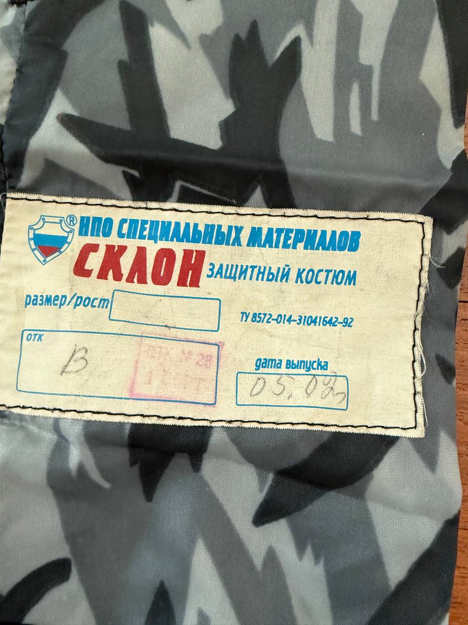 Close-up of a sewn label on the Russian NPO-SM "Sklon" Water-resistant/Windproof Suit – Melted Snow Color featuring Cyrillic text and quality inspection stamps on camouflaged fabric.