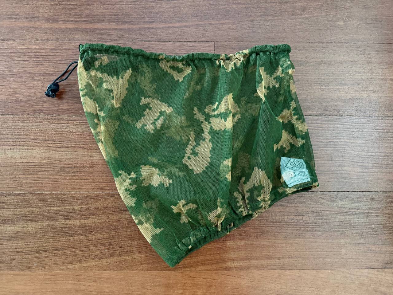 Russian SSO Net Neck Gaiter - Gold Berezka in green and beige camouflage pattern, laid flat on a wooden surface, featuring a drawstring closure and a small fabric tag.