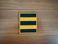 Square embroidered patch with alternating black and orange stripes representing the Russian St. George Ribbon - Military Patch laid on wooden surface.