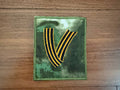 Russian V St. George Ribbon - Military Patch with black and orange stripes on camouflage fabric background, representing Russian surplus military style.