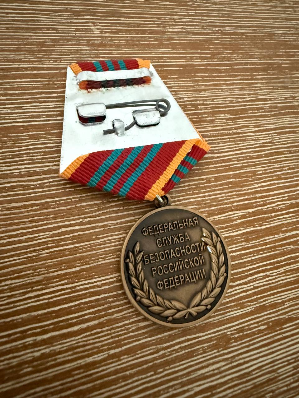 Russian FSB Medal "For Distinction in Military Service" - Replica displayed on a wood grain surface with its pin attachments and a striped red, blue, and yellow ribbon.
