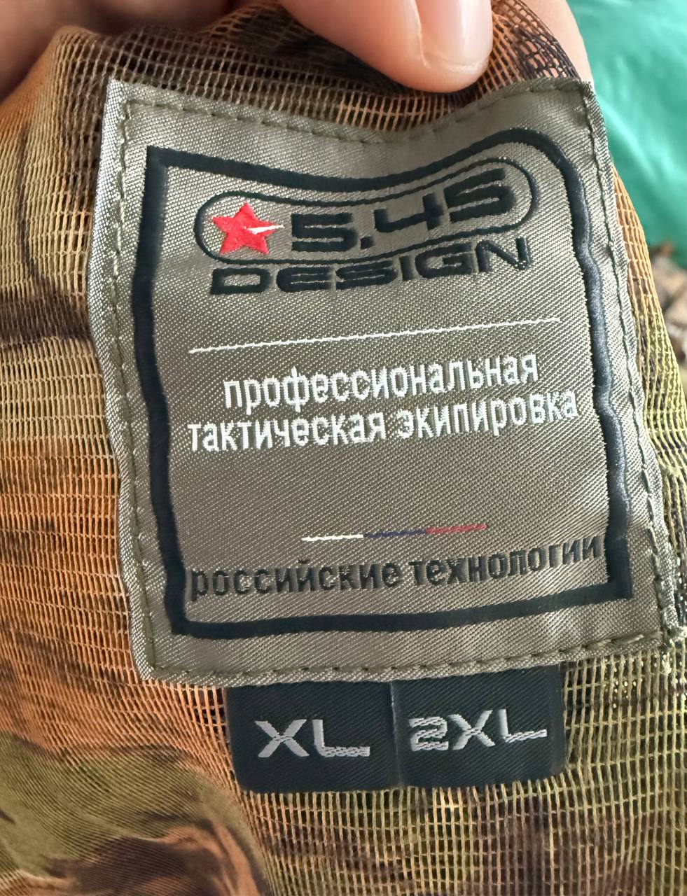 Close-up of a clothing label on the Russian 5.45 Design "Taiga" Leaf Suit – Autumn Camo - Size XL/2XL displaying Russian text and brand logo on camouflage fabric.