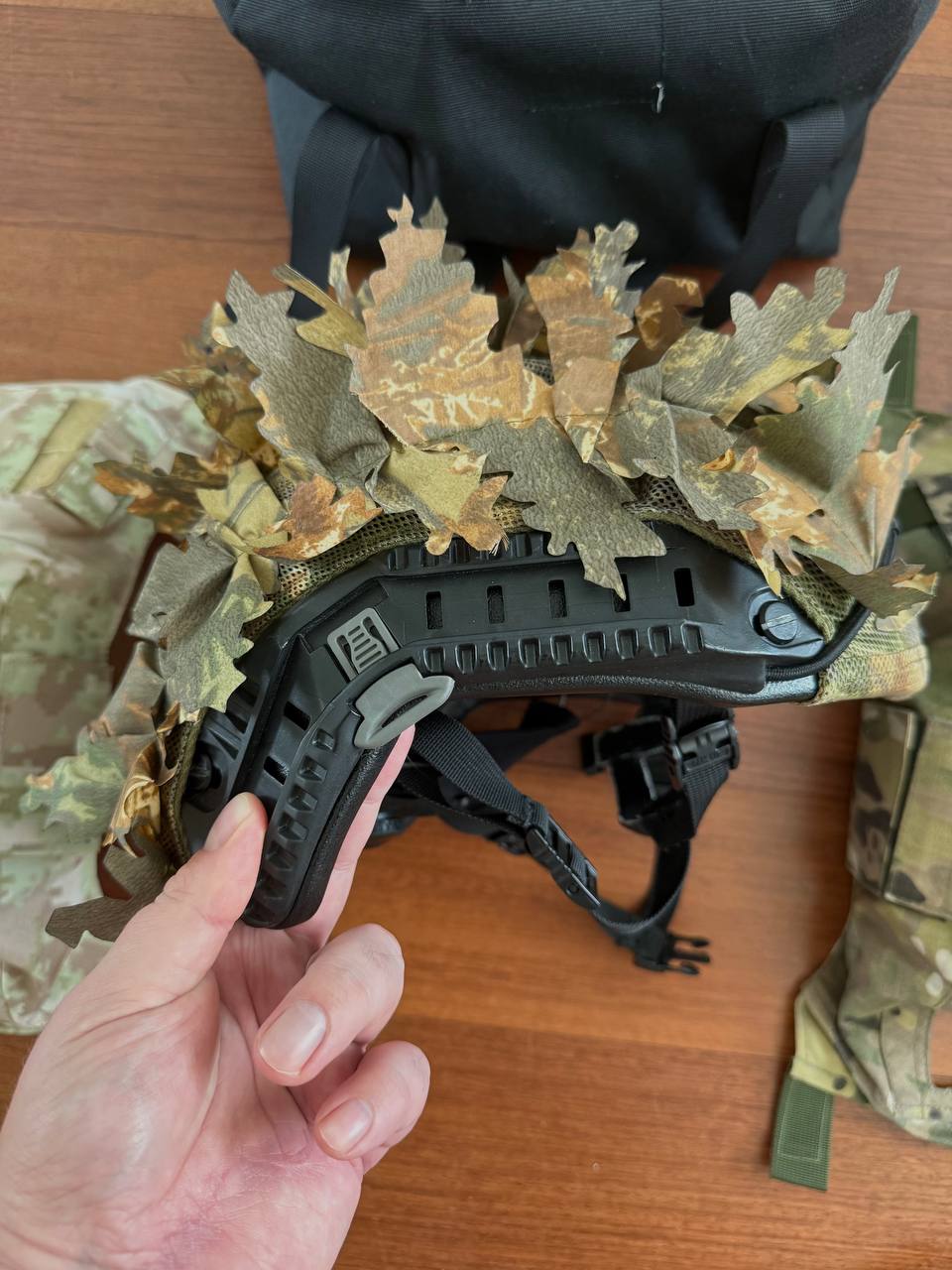 Side view of a Russian LSHZ 1+ Helmet Size 2 with Team Wendy Upgrade and FSB Contract Leaf Cover held by hand, showcasing camouflage leaf attachments and black helmet frame.