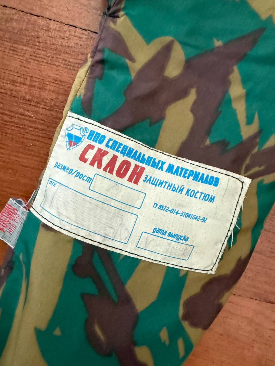 Close-up of the label on a Russian NPO-SM "Sklon" Water-resistant/Windproof Suit – Green Podlesok Color showing text in Russian on camouflage fabric, indicating Russian surplus origin.
