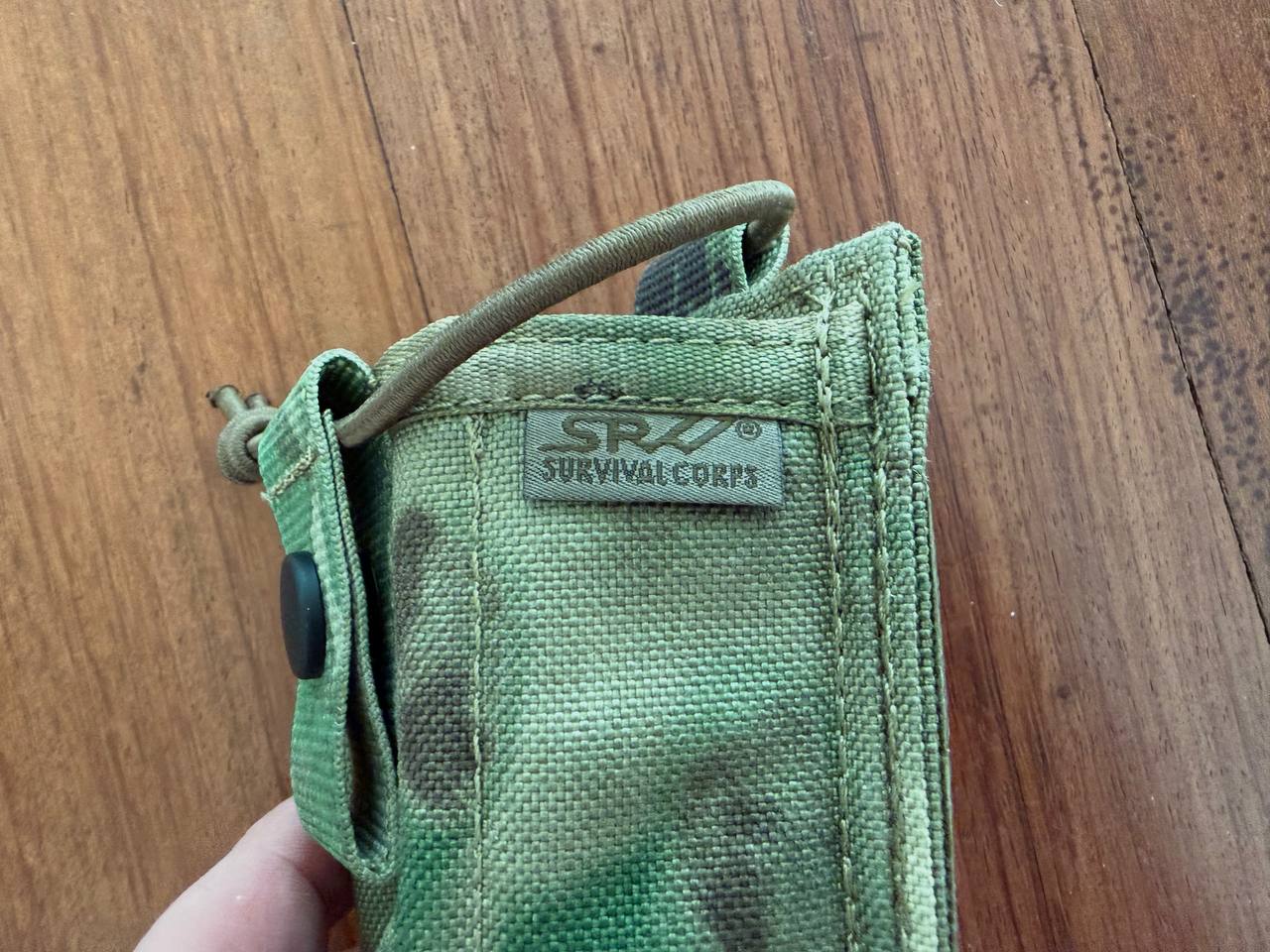 Close-up of a green camo fabric radio pouch with a SRV Survival Corps label held against a wooden background, featuring the Russian SRVV Tactical Radio Pouch - A-TACS FG Camo design.