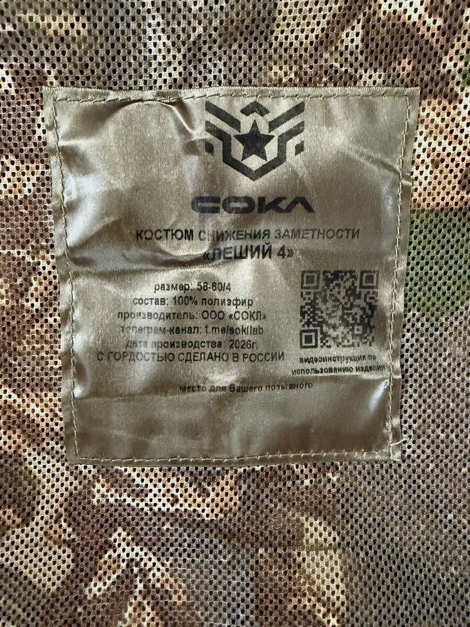 Close-up of a fabric label on a Russian Sokl Leshy 4 Leaf Suit - Autumn Pattern - Size 58-60/4 showing material and manufacturing details with a camouflage mesh background.