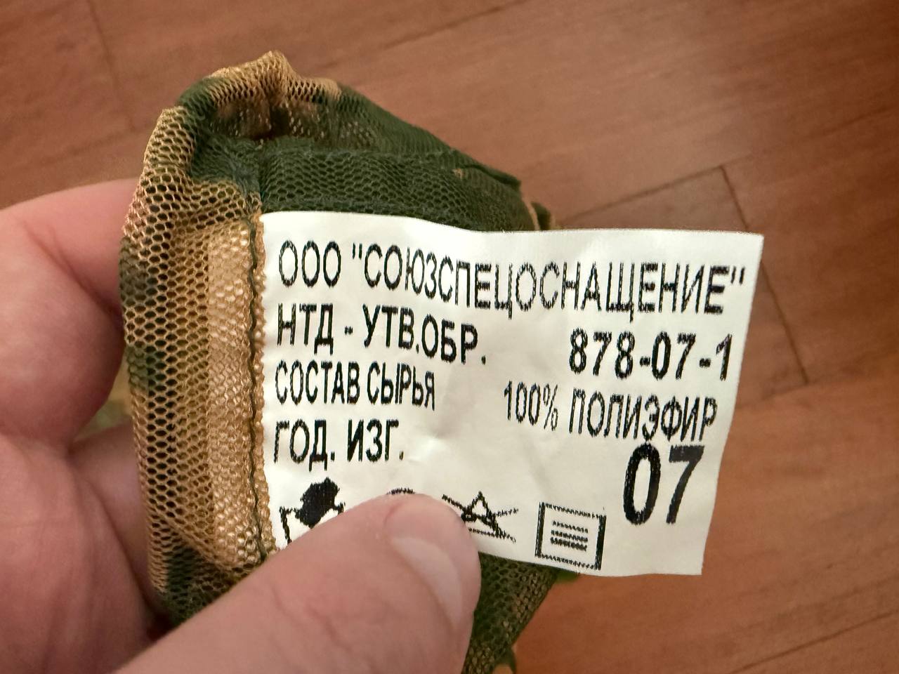 Close-up of a hand holding a care label in Russian on a green and gold mesh fabric, showing material details and manufacturing information for the Russian SSO Net Neck Gaiter - Gold Berezka.