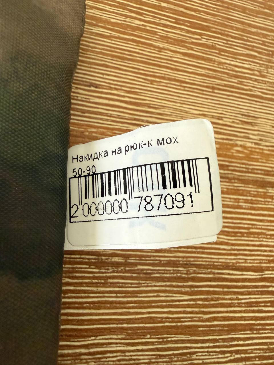 Close-up of a barcode label on a camouflaged fabric item, indicating it is the Russian PROFARMY Waterproof Backpack Cover 30-60L - A-TACS FG Moss, displayed on a wooden surface.