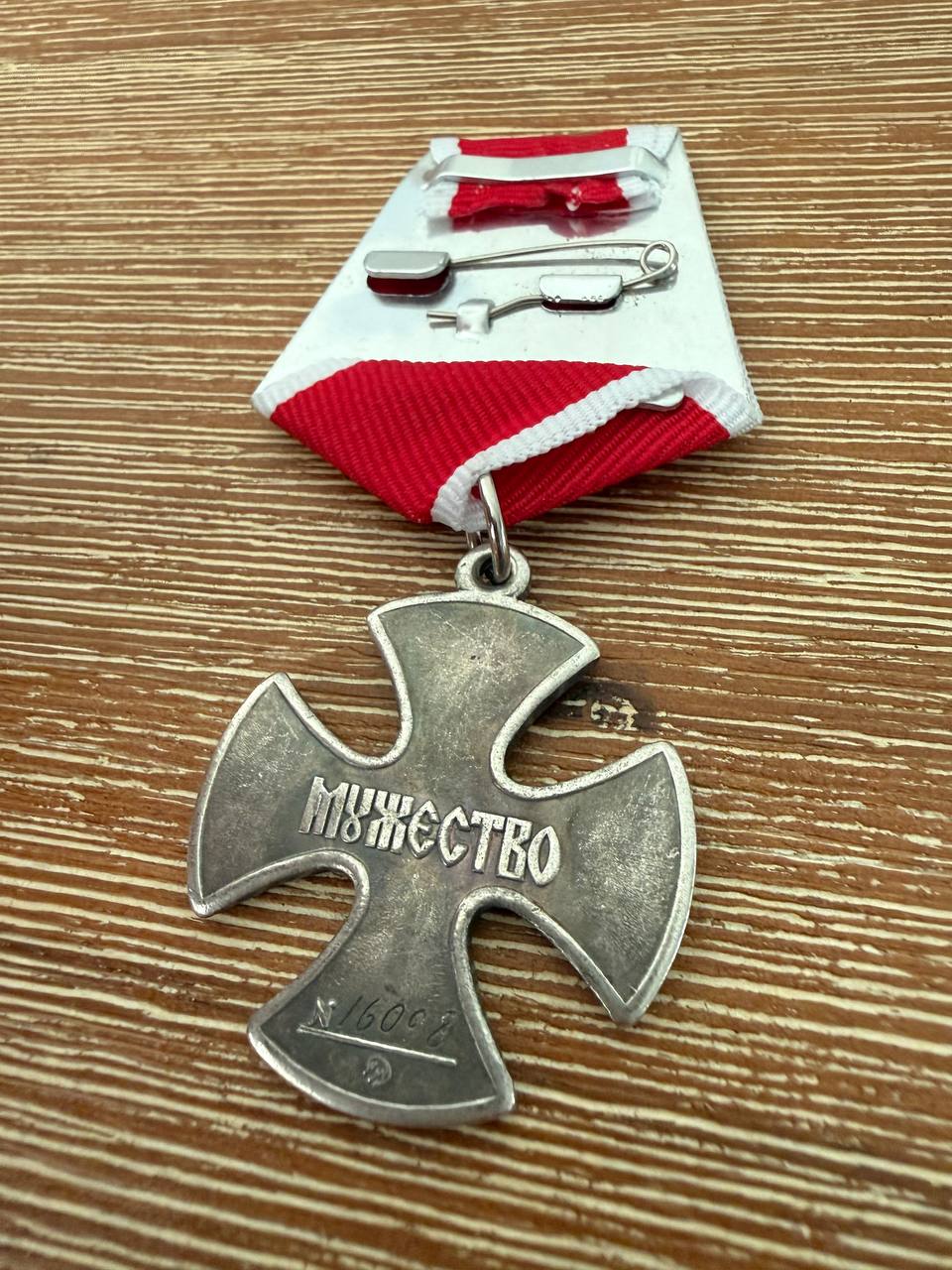 Close-up of a silver cross medal with Russian inscription, attached to a red and white striped ribbon with a pin, identified as the Russian Order of Courage for Military Merit - Replica.