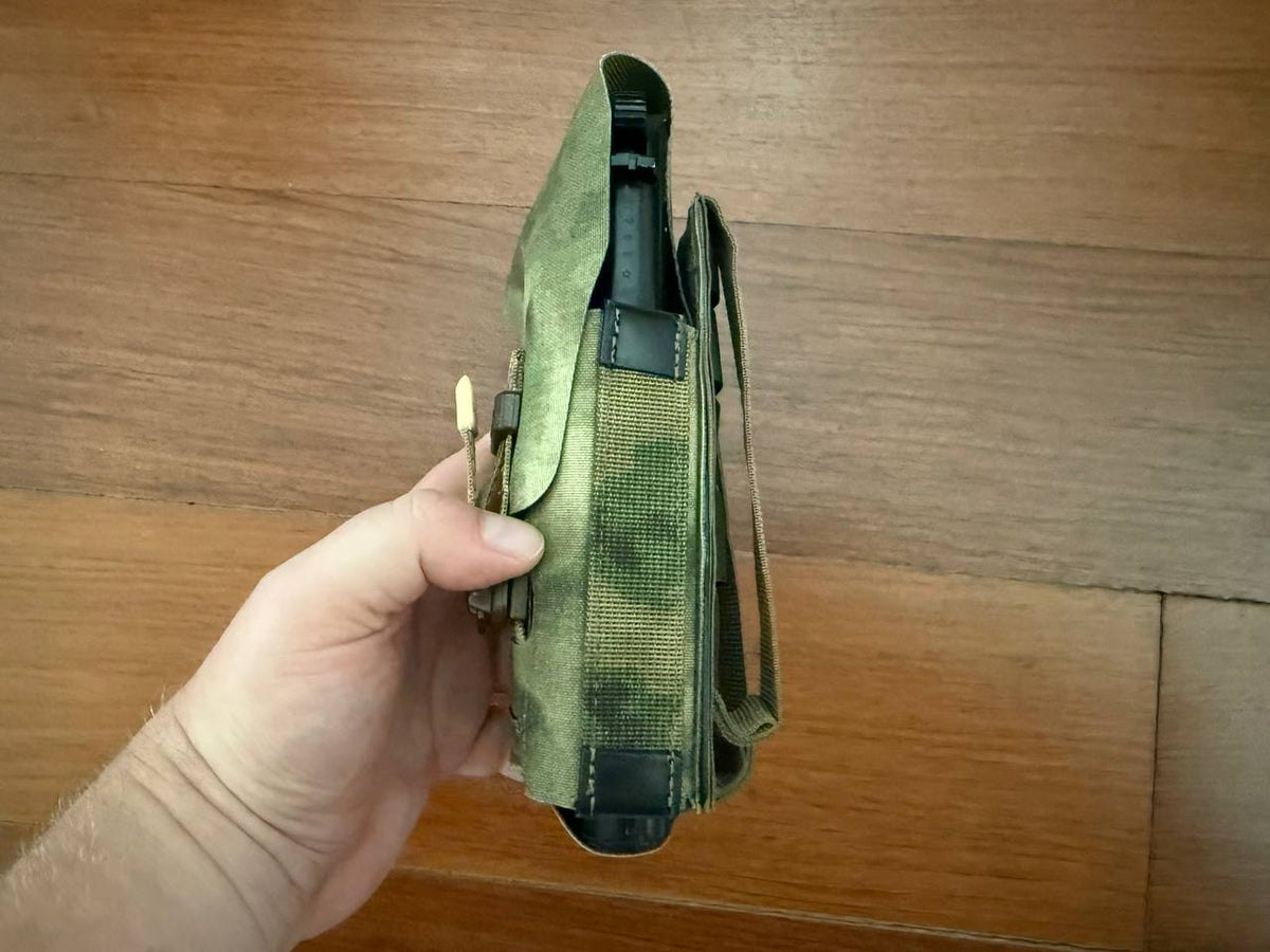 Hand holding a Russian Gear Craft Universal Tactical Holster - A-TACS FG with camouflage pattern and strap visible against a wooden background.