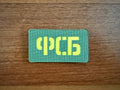 The Russian FSB Unit Patch - Military Badge by Tanki Store is a rectangular green patch with yellow Cyrillic ФСБ, shown on wood. Available in bulk—perfect for collectors seeking unique patches at top value.