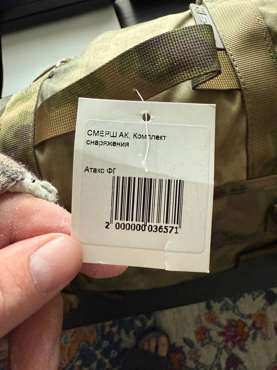 Hand holding a tag on a camouflage bag with a floral carpet in the background
