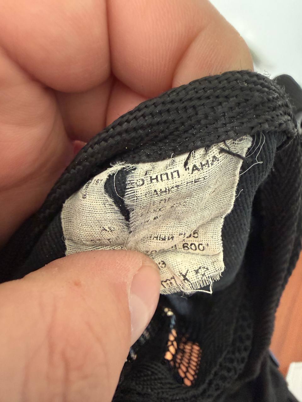 Close-up of a hand holding the interior fabric label of the Russian ANA - "Gorod-2" Tactical Vest – Black, showing worn and faded printed text on the tag.