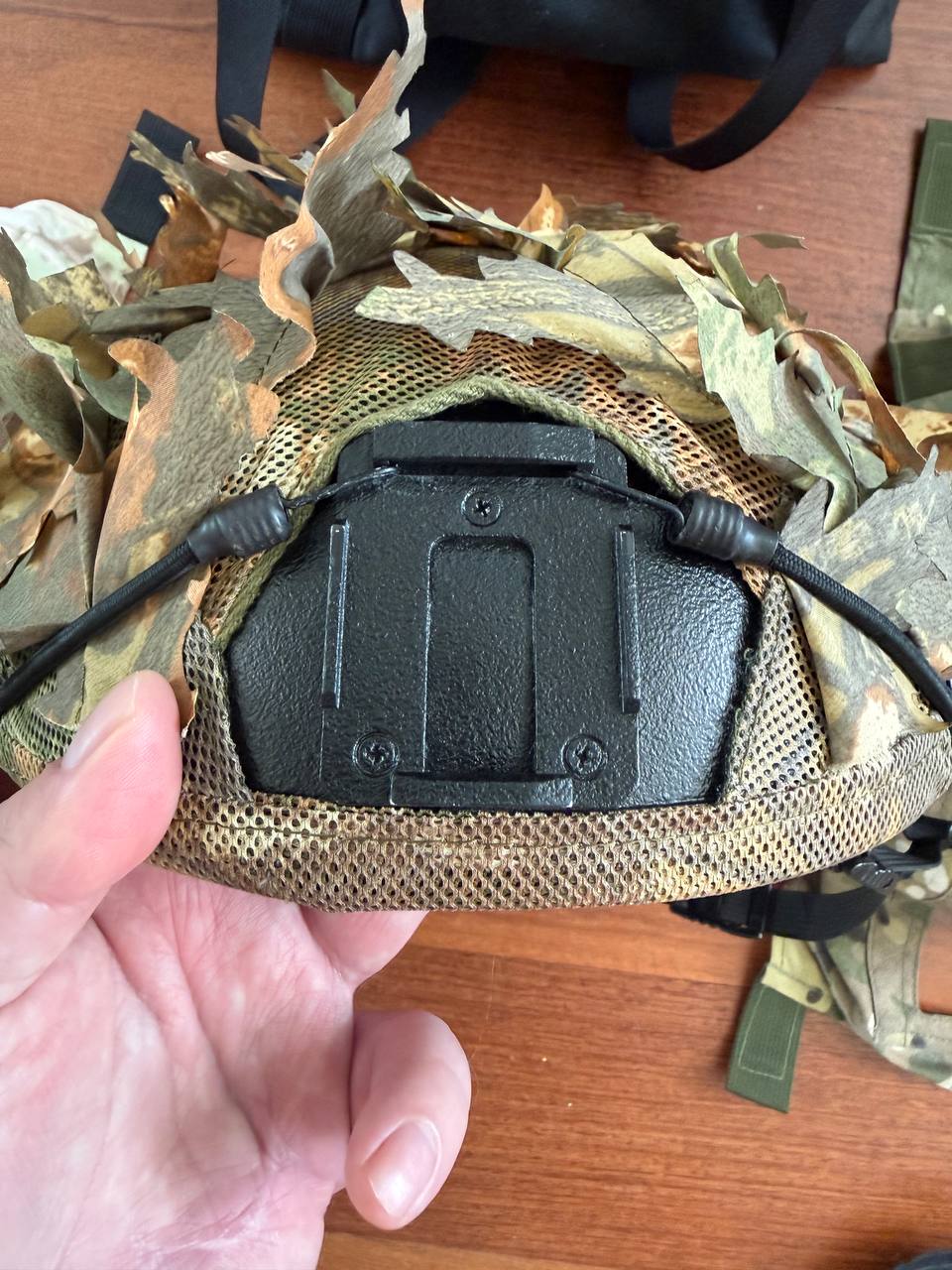 Close-up of a hand holding the underside of a Russian LSHZ 1+ Helmet Size 2 + Team Wendy Upgrade + FSB Contract Leaf Cover featuring camouflage leaf details and black hardware components.