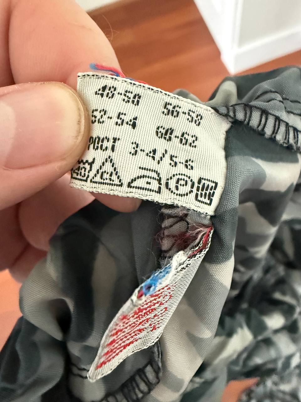 Close-up of a clothing care label on a camouflaged fabric piece, showing washing and ironing instructions for the Russian NPO-SM "Sklon" Water-resistant/Windproof Suit – Melted Snow Color.