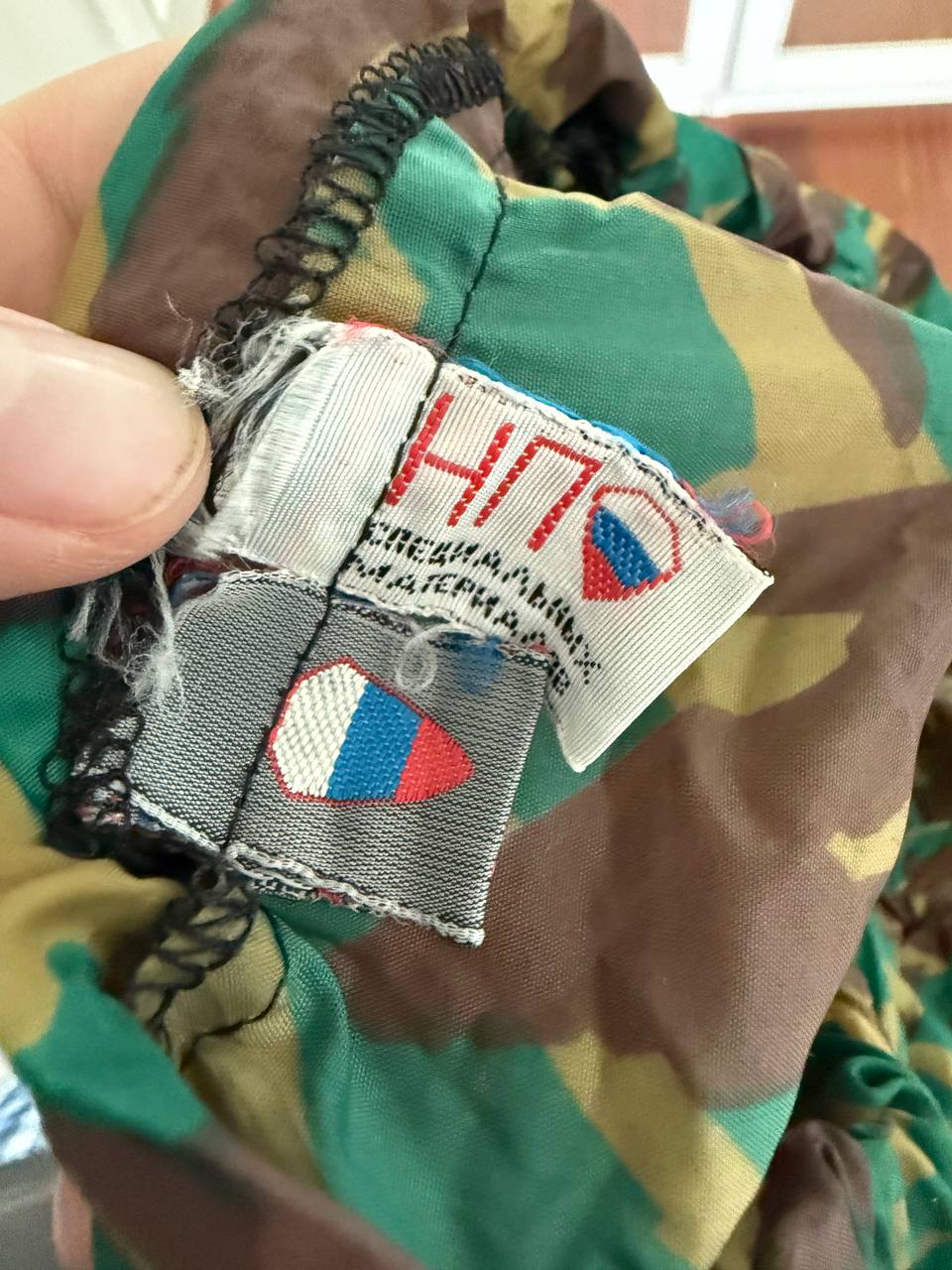 Close-up view of a worn fabric label featuring Russian insignia on camouflage fabric of the Russian NPO-SM "Sklon" Water-resistant/Windproof Suit – Green Podlesok Color.