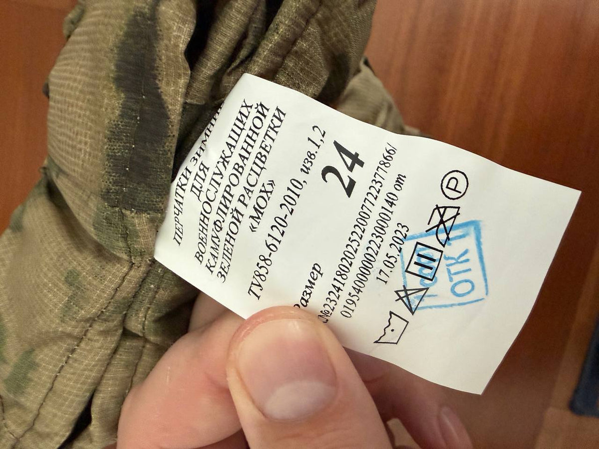 Close-up of a hand holding a tag with size 24 on a pair of Russian Winter Tactical Gloves – A-TACS FG / Moss - Size 24, showing care symbols and manufacturer details.