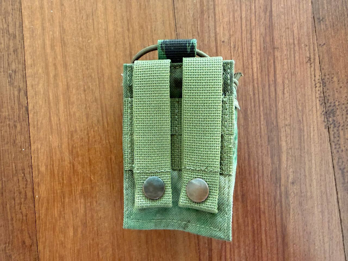 Russian SRVV Tactical Radio Pouch - A-TACS FG Camo shown from the back with dual snap-button straps for secure attachment on a wooden surface.