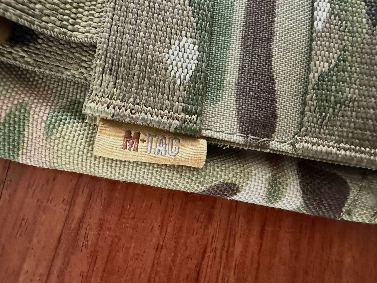 Close-up of the textured fabric and stitching detail on a Ukrainian M-Tac Vest - Multicam showing the tag with the M-Tac logo on a wooden surface.