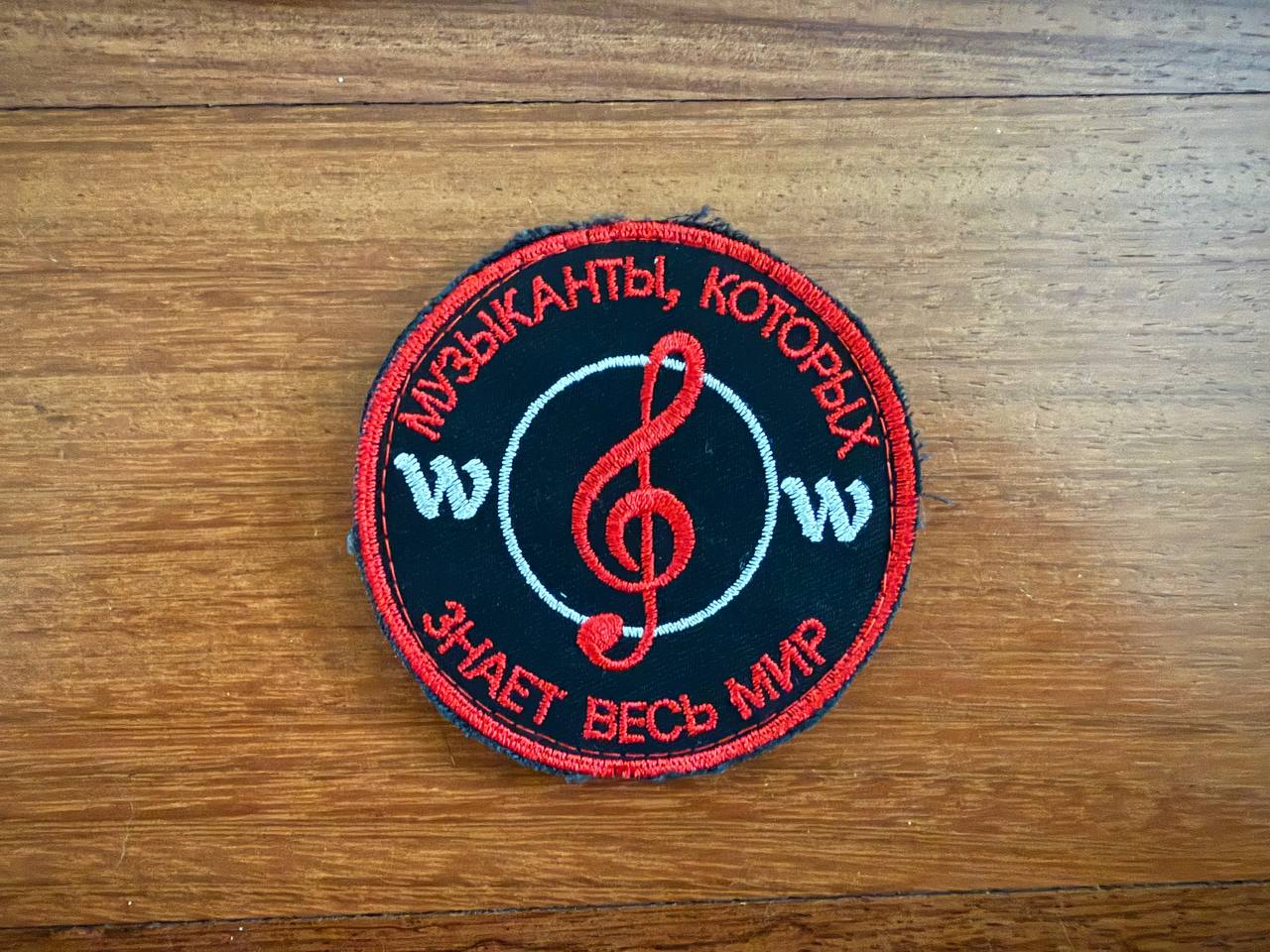 A round black and red PMC Wagner Badge patch from Tanki Store, featuring a treble clef and Russian text Музыканты, которых знает весь мир stitched in red, displayed on a wooden surface. Ideal for military patch collectors.