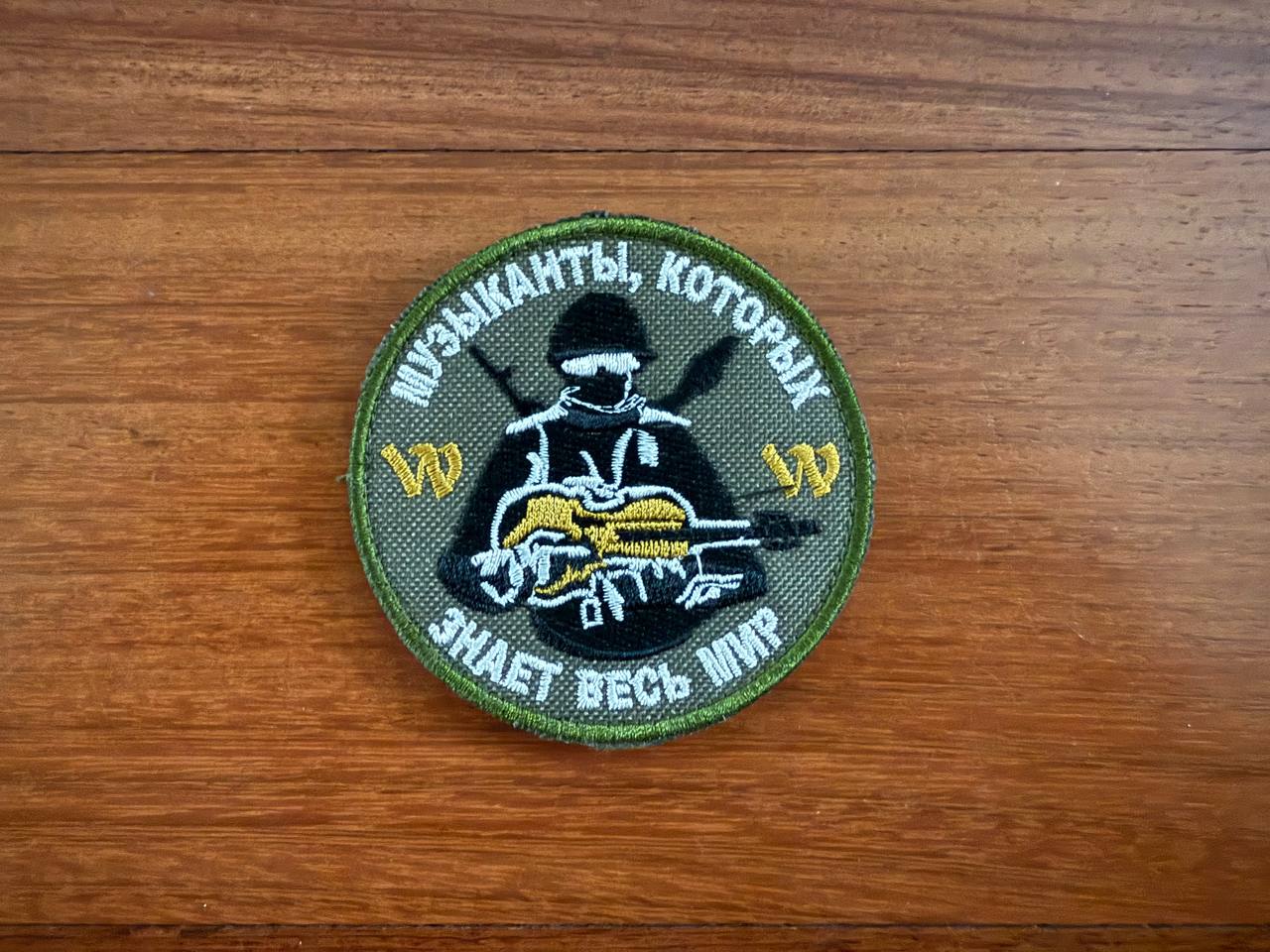 The PMC Wagner Badge - Russian Army Military Patch from Tanki Store features a soldier with a guitar, Russian text, “1/20” in yellow, and a green border—ideal for collectors seeking quality patches at the best price.